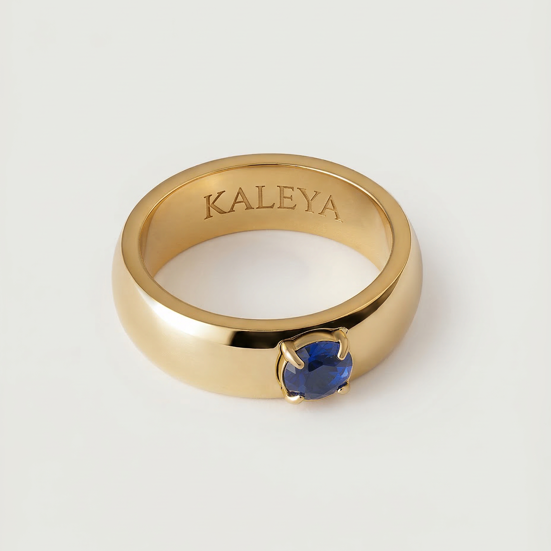 A gold-colored ring with a blue cubic zirconia gemstone in the center, displayed against a white background.