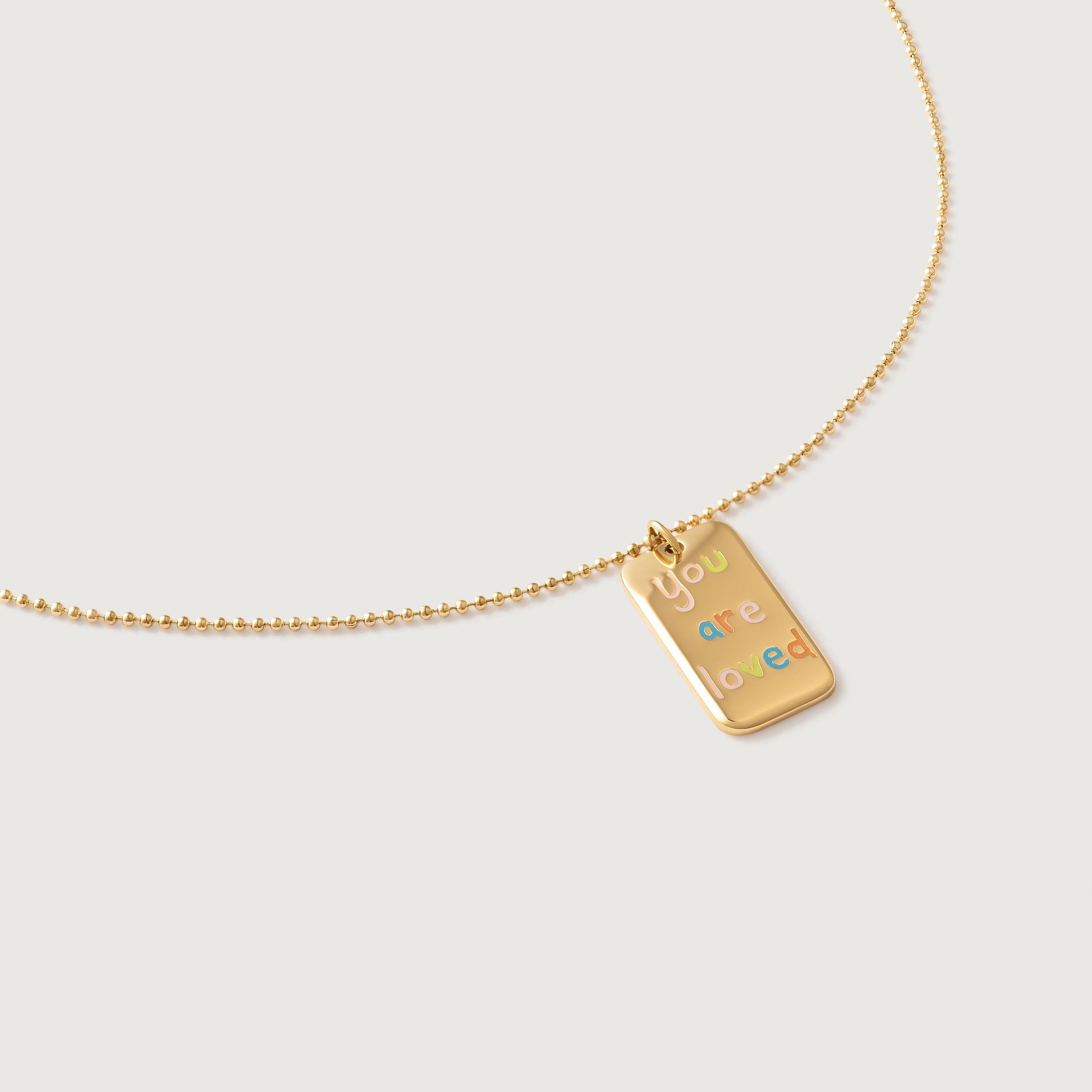 A gold-plated necklace with a polished rectangular tag pendant featuring the phrase 'you are loved' in rainbow enamel letters, attached to a ball chain.