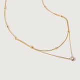 A gold-colored necklace with two layers, featuring tiny spheres on the upper strand and a crystal pendant on the lower chain, displayed against a white background.