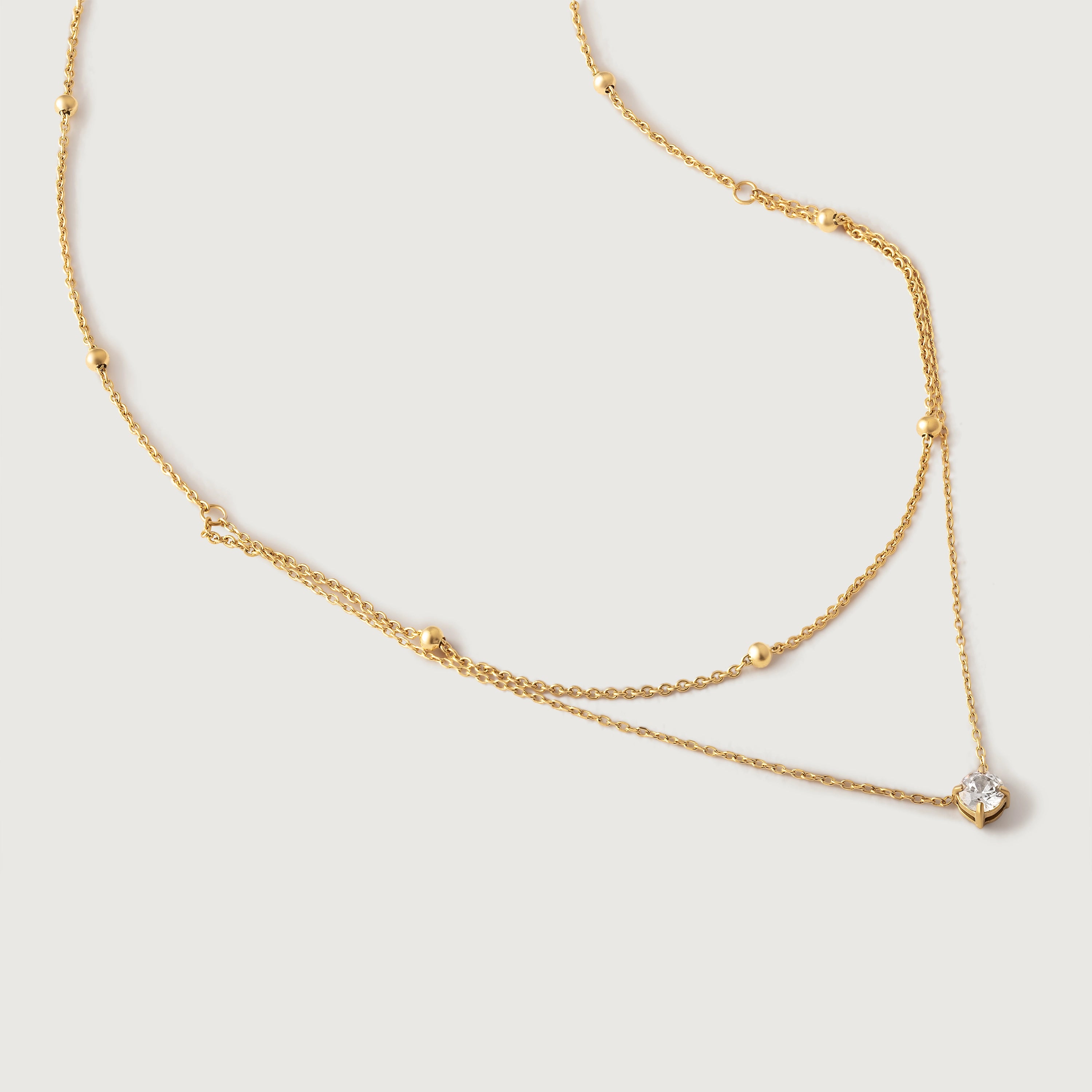 A gold-colored necklace with two layers, featuring tiny spheres on the upper strand and a crystal pendant on the lower chain, displayed against a white background.