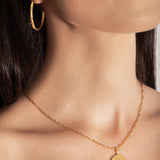 model wearing a gold-colored necklace with a radially textured medallion pendant hanging from a twisted rope chain.