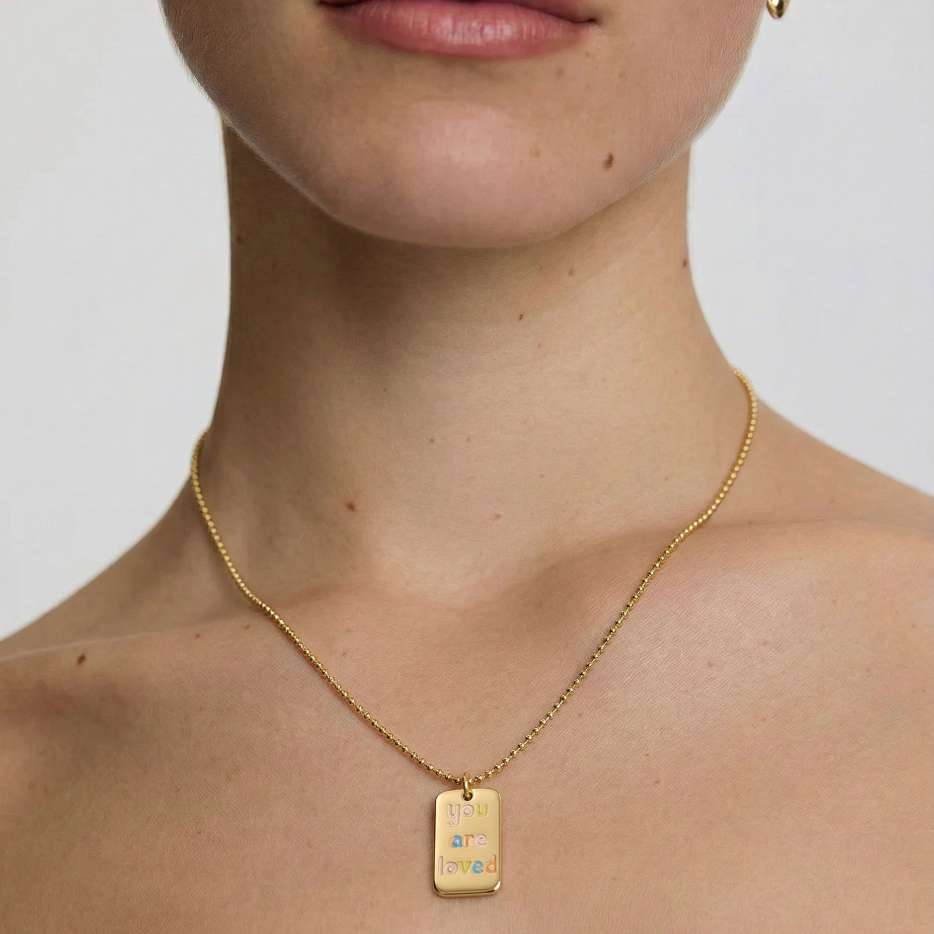 model wearing a gold-plated necklace with a polished rectangular tag pendant featuring the phrase 'you are loved' in rainbow enamel letters, attached to a ball chain.
