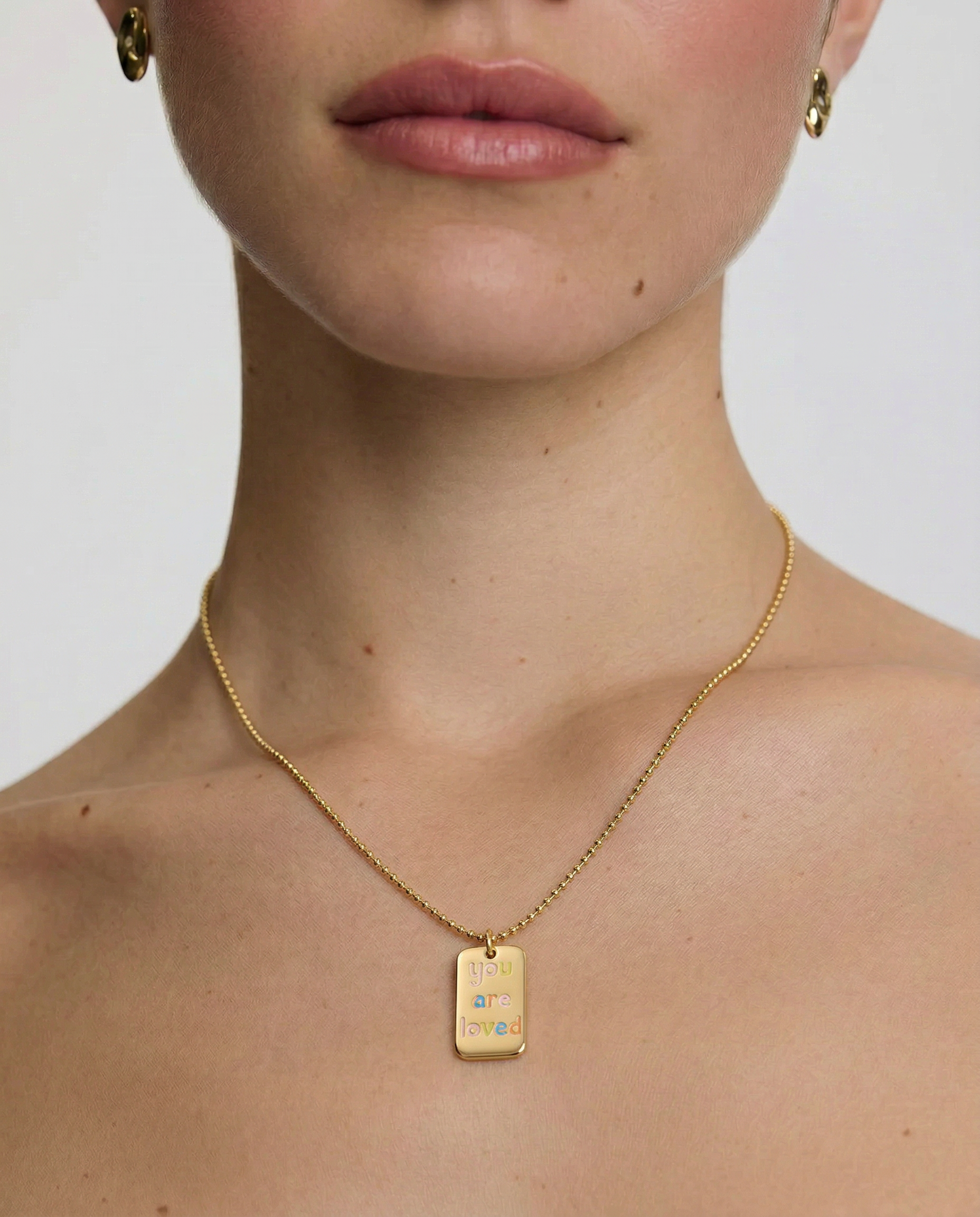 model wearing a gold-plated necklace with a polished rectangular tag pendant featuring the phrase 'you are loved' in rainbow enamel letters, attached to a ball chain.
