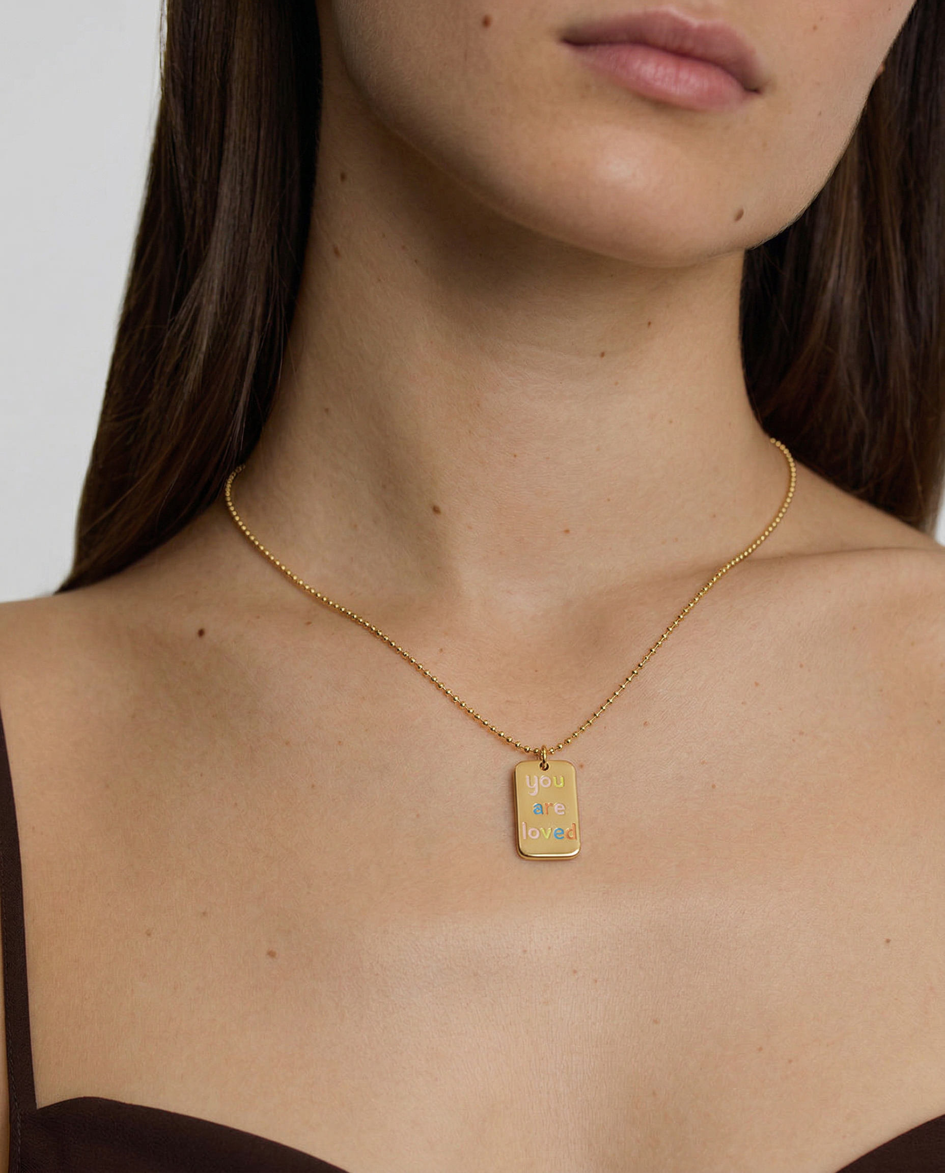 model wearing a gold plated necklace with a pendant saying "you are loved" in rainbow letters