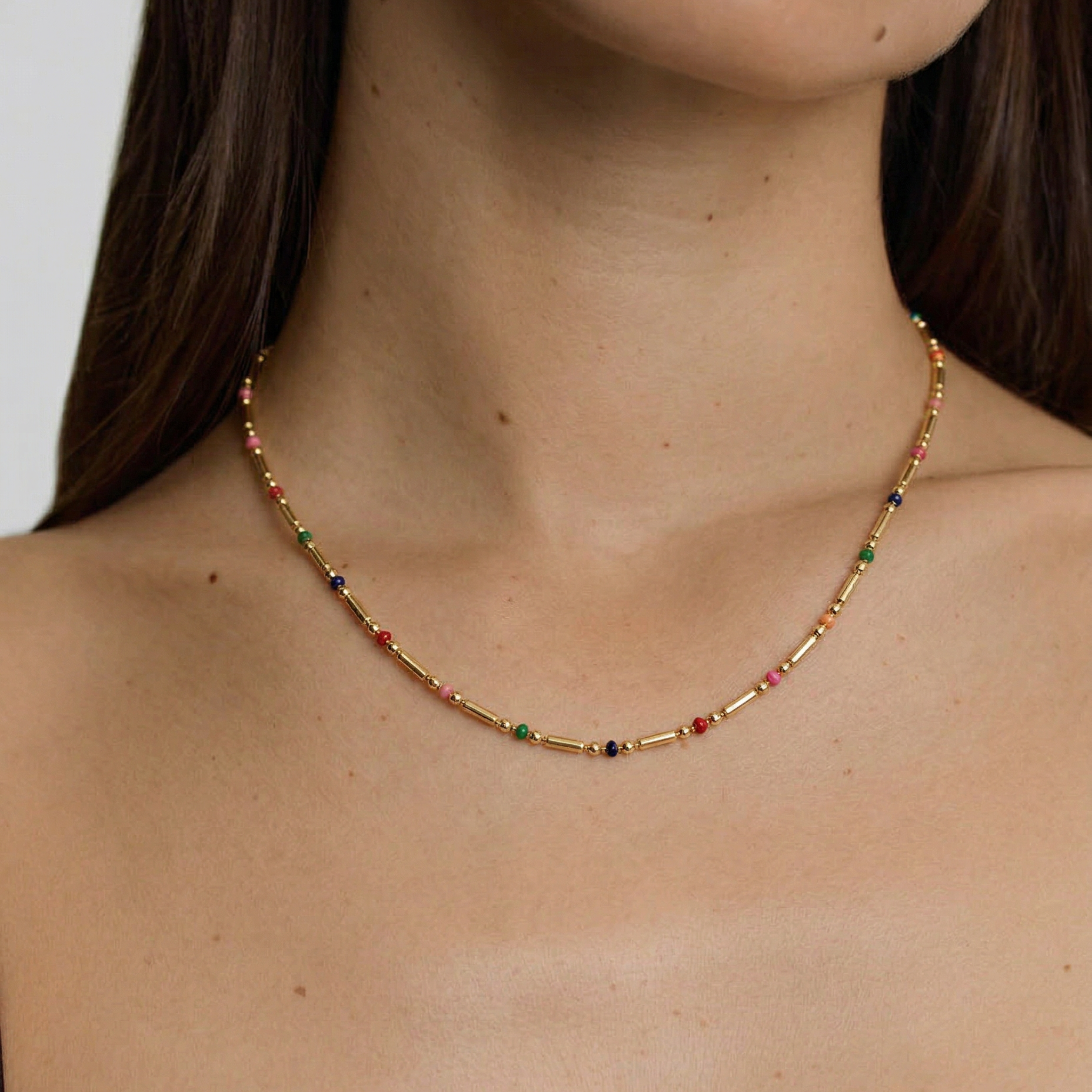 female model wearing a gold-plated necklace with multicolored beads in red, pink, blue, and green, arranged in a alternating pattern between golden bars.