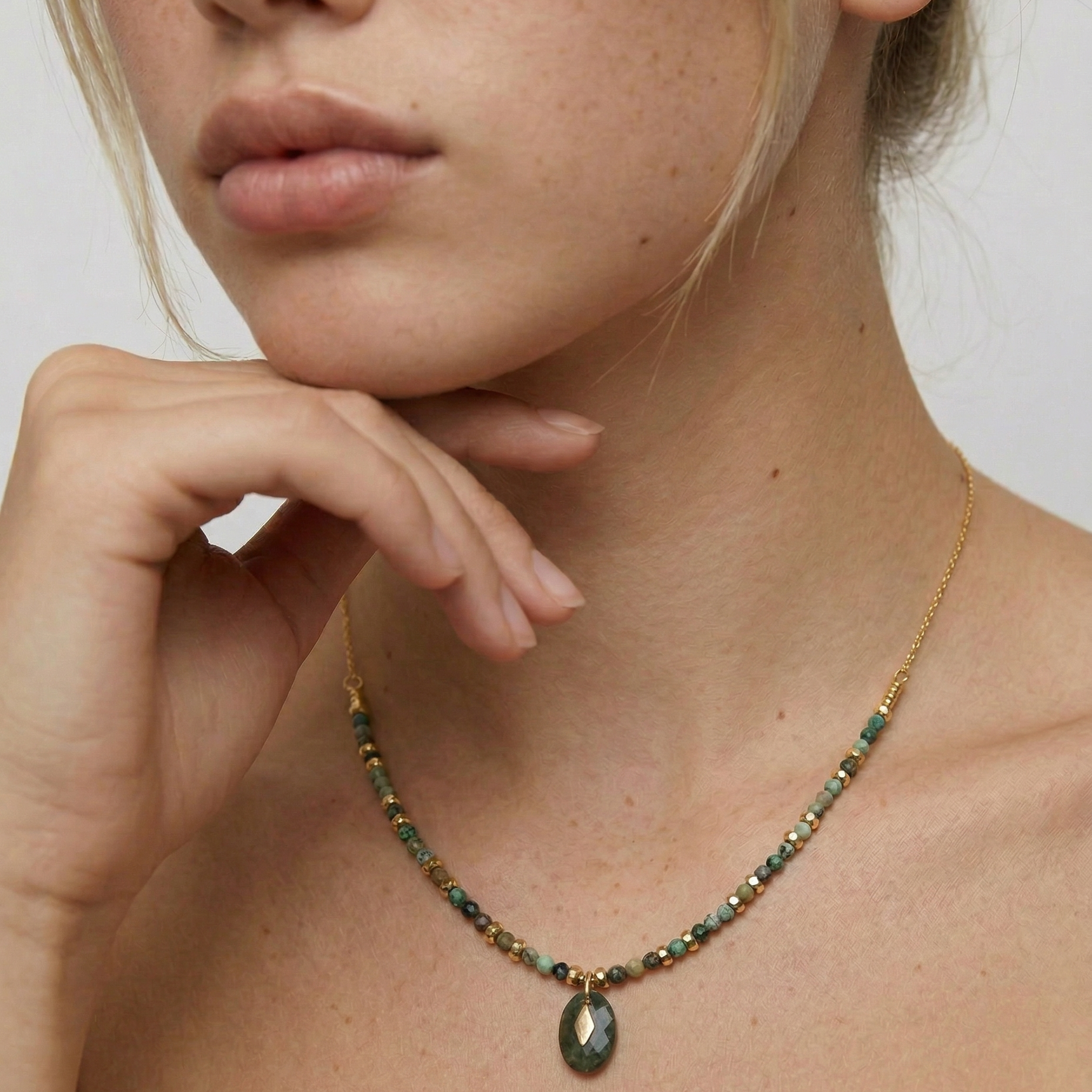Close-up of a person wearing a gold-toned necklace with natural African turquoise beads in shades of sage, moss, and earth tones, featuring a smooth oval pendant.