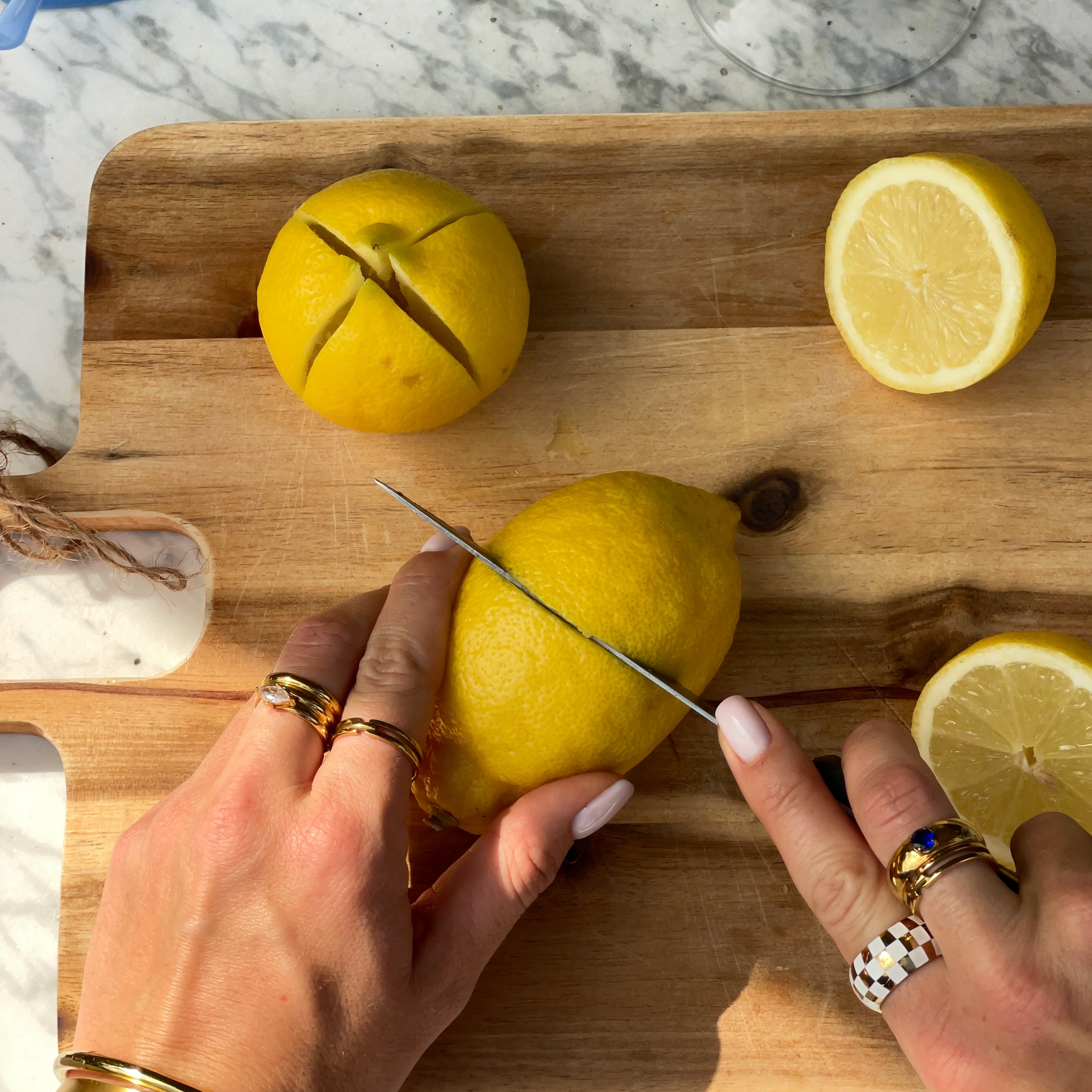Person wearing gold plated jewellery cutting a lemon on a wooden cutting board with a knife.